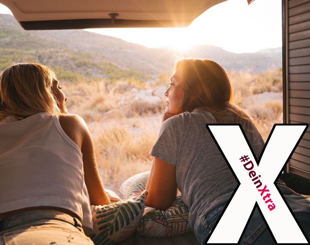 #DeinXtra | Bild zeigt zwei Frauen die zufrieden aus ihrem Camper auf eine sonnige Landschaft blicken. | © AdobeStock: ManuPadilla